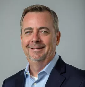 A professional portrait of a man wearing a navy blue blazer and a light blue collared shirt. The background is a neutral gray, emphasizing the formal attire. The image conveys a corporate or business setting.